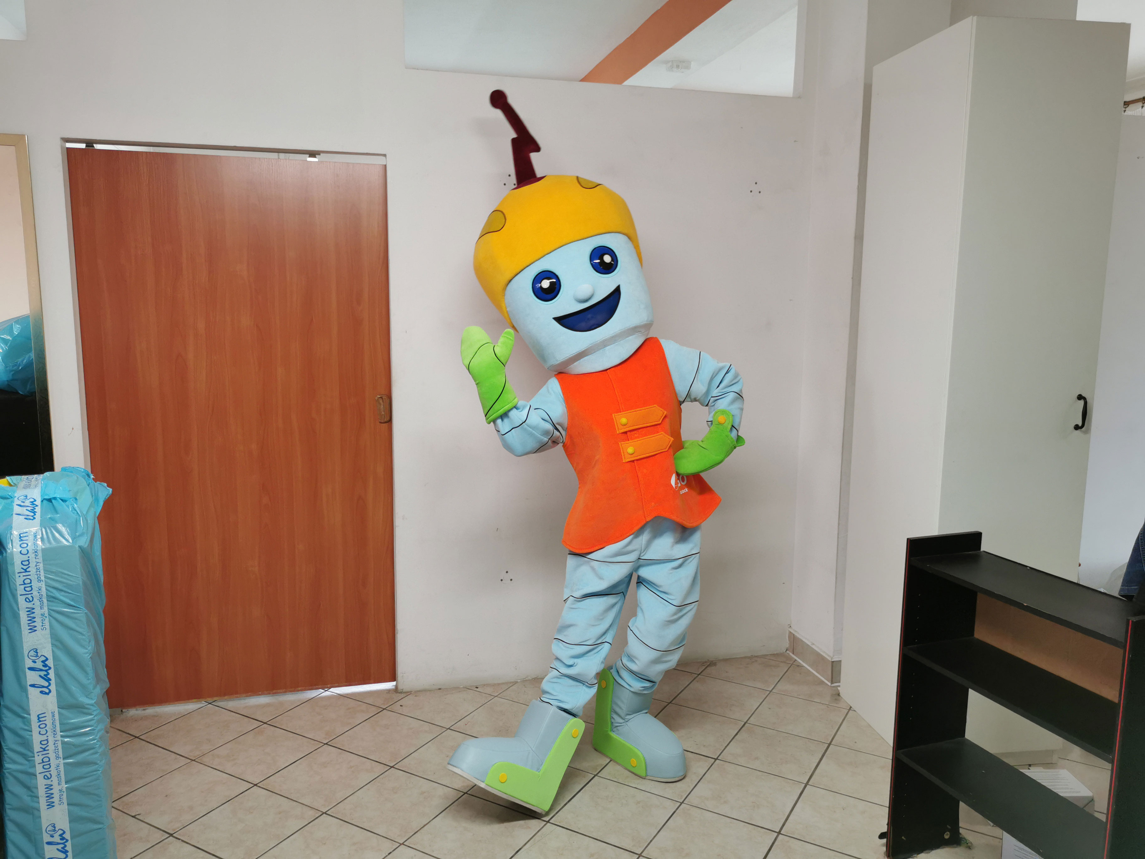 Na fotografii widnieje strój reklamowy w postaci ludzika pozujący do zdjęcia w siedzibie firmy Elabika